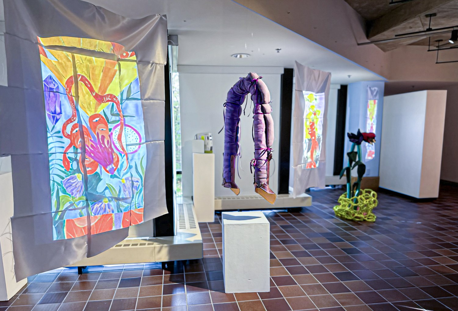 Installation image of art by Ivonne Yañez. Along a diagonal wall, moving image is projected onto fabric tapestries, alongside hanging soft sculptures in shiny purple silk.