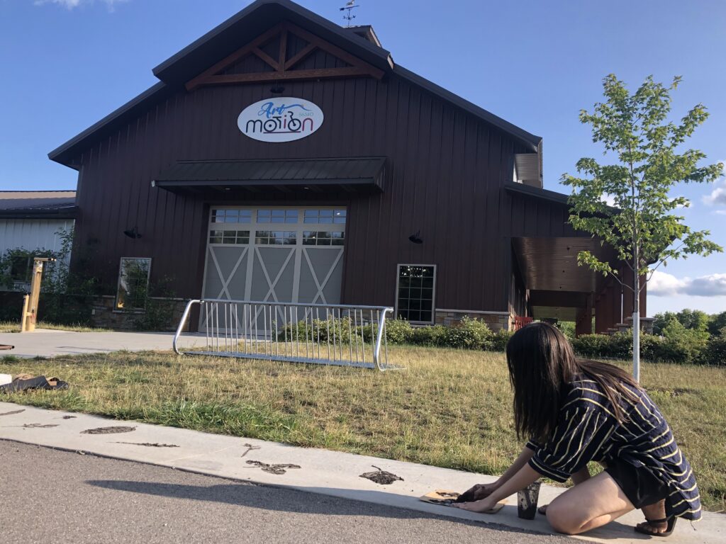 an image showing Malini crouching in the foreground applying mud to her stencils, her hair covers her face. in the background you can see the Boho Cafe and a sign that reads, "Art in Motion."