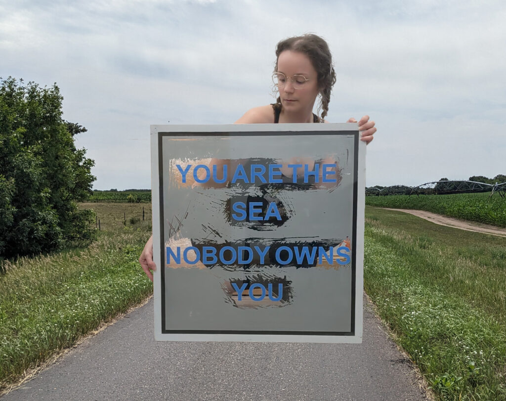 A digital collage including an image of the Lake Wobegon Trail and a larger-than-life sized Liz holding a painting that reads, "You are the sea, nobody owns you."